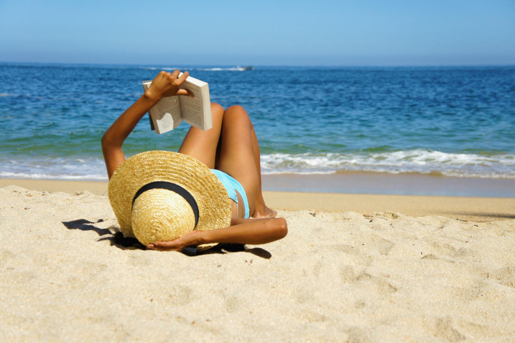 Woman lying on a beach wearing a sunhat and reading a book.