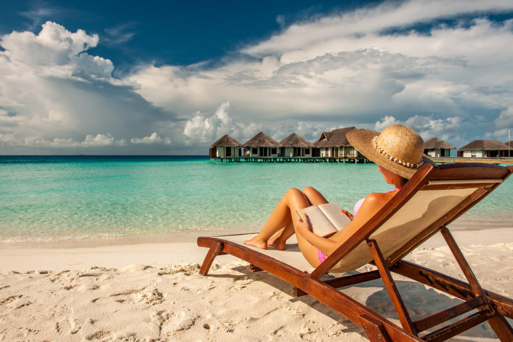 Woman sitting on a beach lounge chair, looking over the calm ocean, reading a book.